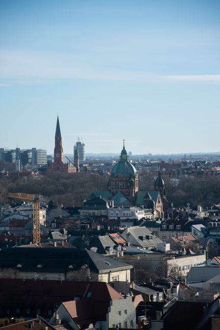 A panoramic view of a cityscape with a mix of historic and modern architecture, including prominent church spires and domed rooftops, under a clear blue sky. In the foreground, rooftops of residential